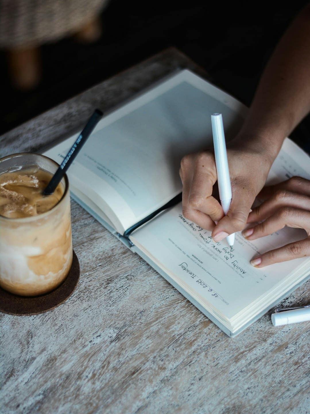 Hände schreiben mit weißem Stift in ein Notizbuch mit Eiskaffee.