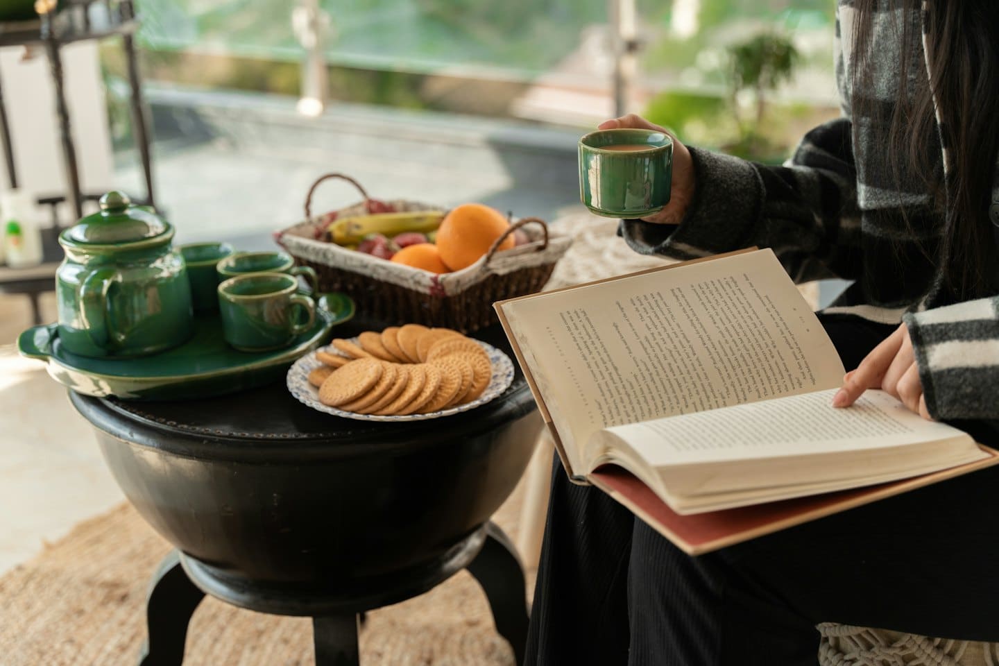 Eine Person liest ein Buch am Fenster bei Tee und Obst.